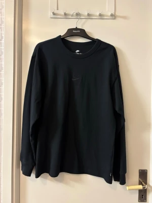 Svart långärmad sweatshirt från Nike - Svart långärmad sweatshirt från Nike med diskret broderad logga på bröstet. Tröjan har rund halsringning och är tillverkad i mjukt bomullsmaterial. Perfekt för en avslappnad och sportig look.