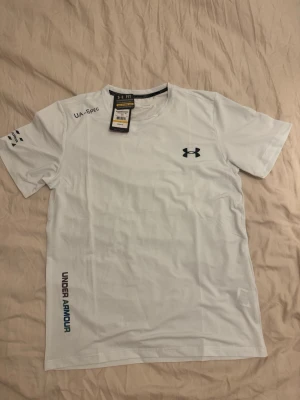 Vit Under Armour t-shirt - Säljer en vit t-shirt från Under Armour med logga på bröstet och tryck på sidan. T-shirten har rund hals och korta ärmar, perfekt för dig som gillar sportiga och stilrena plagg. Materialet är syntet och känns lätt och bekvämt.