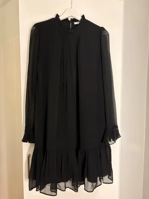Svart volangklänning med lång ärm - Säljer en svart kortklänning med transparenta långärmar och volangdetaljer vid ärmslut och nederkant. Klänningen har hög hals och ett lätt, flödande tyg som ger en luftig känsla. Perfekt för dig som gillar stilrena och trendiga plagg.