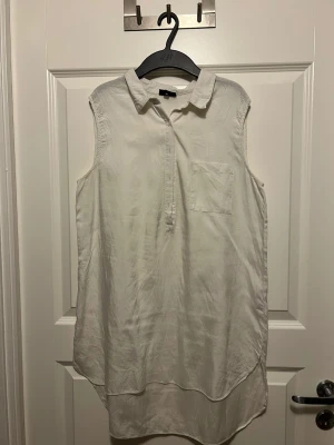 Vit ärmlös blus från KappAhl - Stilren vit ärmlös blus från KappAhl med klassisk krage och bröstficka. Blusen har en avslappnad passform och knappar framtill. Perfekt till både jeans och kjol för en fräsch look. Materialet känns mjukt och svalt, troligen bomull eller viskos.