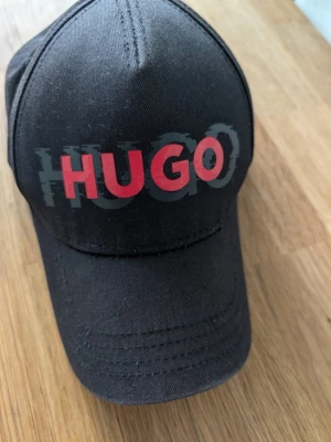 Svart keps från Hugo Boss - Snygg svart keps från Hugo Boss med stor röd och grå HUGO-logga framtill. Klassisk böjd skärm och justerbar passform bak. Tillverkad i bomull för en bekväm känsla och stilren look. Perfekt accessoar för att lyfta din outfit.