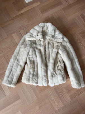 Beige fuskpälsjacka från Milla - Snygg beige ljus fuskpälsjacka från Milla i storlek S. Jackan har en fluffig krage, dragkedja framtill och är tillverkad i mjukt fuskpälsmaterial. Perfekt för att hålla värmen under kalla dagar och ger en lyxig känsla till din outfit.