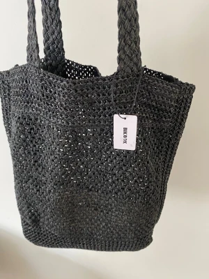 Svart stickad tote från Bik Bok strandväska - Snygg svart stickad tote bag från Bik Bok med flätade handtag. Väskan är gjord i ett grovt, öppet stickat material som ger en avslappnad och trendig look. Perfekt att slänga över axeln när du ska ut på stan eller till stranden.