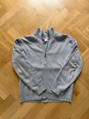 Grå ziptröja från Lindex - En stilren grå ziptröja från Lindex med hög krage och ribbade muddar. Tröjan är tillverkad i mjukt stickat material och har en enkel design som passar perfekt till jeans eller joggers.