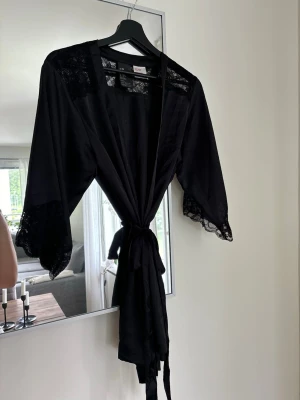 Svart kimonomorgonrock med spets H&M - Snygg svart morgonrock i kimonomodell från H&M. Tillverkad i ett tunt, mjukt tyg med vackra spetsdetaljer på axlar och ärmslut. Knytband i midjan för en skön passform. Perfekt för chill dagar hemma.