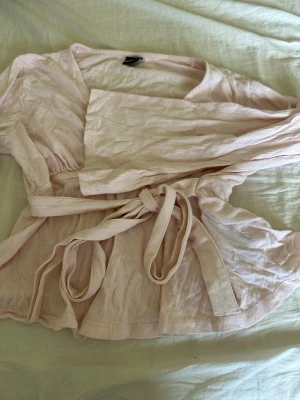 Ljusrosa omlottblus Gina Tricot - Supersöt ljusrosa omlottblus från Gina Tricot med knytband i midjan och lätt rynkade detaljer. Blusen har lång ärm och är gjord i ett tunt, mjukt tyg som känns luftigt och skönt. Perfekt att styla med jeans eller kjol för en fräsch look.
