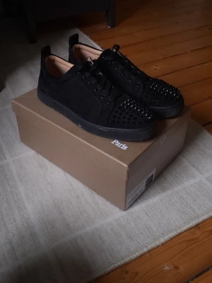Svarta Louboutin sneakers med nitar - Svarta sneakers från Christian Louboutin i mocka med coola svarta nitar på tån och svarta skosnören. Snygg och edgy design med låg profil och klassisk röd innersula. Perfekta för dig som vill sticka ut med din stil.