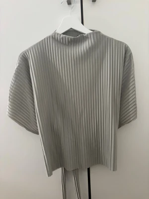 Plisserad grön blus med knytband - Stilren grön plisserad blus med korta ärmar och knytband i midjan. Blusen är tillverkad i ett glansigt, lätt material som ger en modern look. Perfekt för dig som gillar minimalistisk stil.