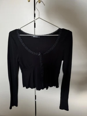 Svart långärmad topp från Gina  - Svart långärmad topp från Gina tricot  med spetskant längs ringningen och knäppning framtill. Toppen har en liten rosett vid halsen och vågig, croppad nederkant. Perfekt för en trendig och stilren look.