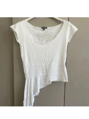 Vit asymmetrisk topp från Sisters - Vit topp från Sisters med korta ärmar, asymmetrisk nederkant och en broderad fjäril i spets vid halsringningen. Tunt och mjukt material som känns skönt mot huden. Perfekt för dig som gillar unika detaljer och en lite annorlunda look.