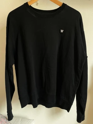 Svart sweatshirt från Wood Wood - Snygg svart sweatshirt från Wood Wood med rund halsringning och diskret logotyp-patch på bröstet. Tröjan har en klassisk passform och är tillverkad i mjuk bomull. Perfekt för en stilren och avslappnad look. Passar M och S 