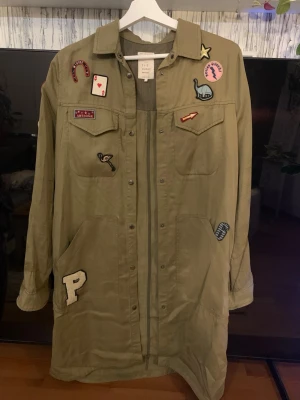 Olivgrön overshirt med patches - Säljer en olivgrön overshirt från One Teaspoon med coola broderade patches som stjärna, dinosaurie, läppstift och bokstaven P. Lång modell med stora fickor fram, tryckknappar och klassisk krage. Perfekt för dig som vill sticka ut lite extra.