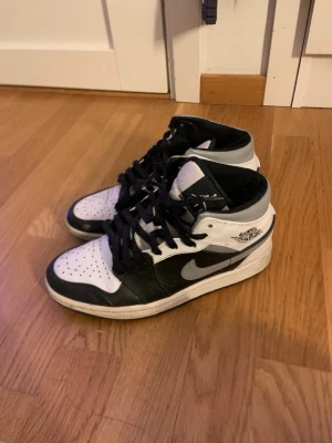 Nike Air Jordan 1 Mid svart/vit - Snygga Nike Air Jordan 1 Mid sneakers i svart och vitt med klassisk Swoosh-logga på sidan och Air Jordan Wings-märke vid ankeln. Skorna har svart snörning, perforerad tåbox och är tillverkade i läder och textil. Perfekt för dig som gillar streetwear och ikoniska sneakers. De är köpta på plick. Någon fläck som du kan se på bilden. Hälen inne är lite sämdrig (se bild) men de funkar annars. 