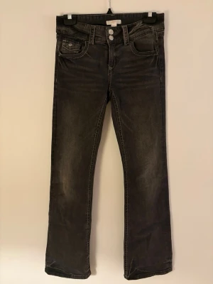 Svarta bootcut jeans med dubbla knappar - Snygga svarta bootcut jeans med dubbla knappar i midjan och klassiska fem fickor. Jeansen har en lätt tvättad look och raka ben med svag utsvängning. Perfekta för en avslappnad och trendig stil.