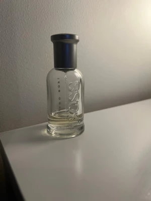 Hugo Boss Bottled parfym - Stilren parfymflaska från Hugo Boss med genomskinligt glas och silverfärgad kork. Flaskan har en rund form och tydlig BOSS-logga ingraverad på framsidan. Perfekt för dig som gillar klassiska och fräscha dofter. Runt 20 ml kvar.