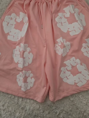 Denim Tears Shorts - Snygga rosa shorts med vita blommönster och resår i midjan med snörning. De har en avslappnad passform och är perfekta för varma dagar. Materialet känns mjukt och bekvämt mot huden.
