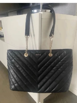 Svart quiltad handväska från DKNY - Snygg svart handväska från DKNY med quiltat mönster i chevronstil. Väskan har guldfärgade kedjedetaljer på axelremmarna och är tillverkad i läderimitation. Perfekt för dig som vill ha en stilren och trendig accessoar.