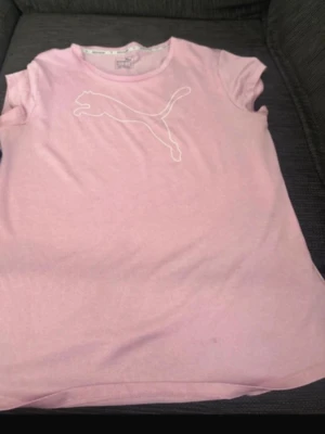 Rosa t-shirt från Puma - Snygg ljusrosa t-shirt från Puma med klassisk logga framtill. T-shirten har korta ärmar och är tillverkad i ett lätt och bekvämt material som andas. Perfekt för en sportig och avslappnad stil.