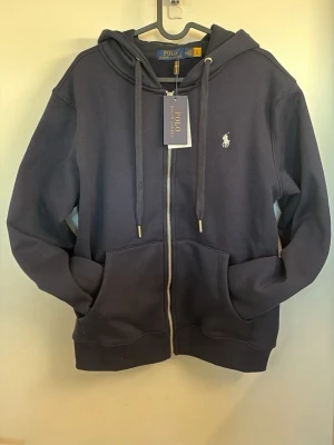 Mörkblå hoodie från Polo Ralph Lauren - Mörkblå hoodie med dragkedja från Polo Ralph Lauren. Klassisk design med huva, dragsnören och två stora fickor framtill. Ikoniska vita loggan broderad på bröstet. Mjuk bomullsblandning som är skön att bära.