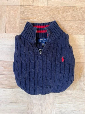 Mörkblå kabelstickad tröja Polo Ralph Lauren - Mörkblå kabelstickad tröja från Polo Ralph Lauren med halv dragkedja och röd broderad logga på bröstet. Tröjan har ribbade muddar och krage samt röd-randigt foder i halsen. Perfekt för dig som gillar klassisk och stilren look.