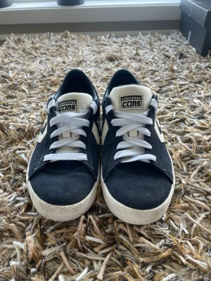Converse CONS svarta sneakers - Svarta Converse CONS sneakers med vit sula och vita detaljer. Klassisk låg modell med vit logga på plösen och sidorna. Ovandel i mocka och textil, gummisula med mönster undertill. Perfekta för en streetwear-look.