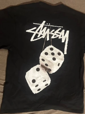 Svart Stüssy t-shirt med tärningsmotiv - Svart t-shirt från Stüssy med stort vitt logotryck och två tärningar på ryggen. Framtill finns en mindre Stüssy-logga på bröstet. Klassisk passform och rund hals. Perfekt för dig som gillar streetwear och vill sticka ut. T-shirten är egentligen storlek S men känns mer som XS