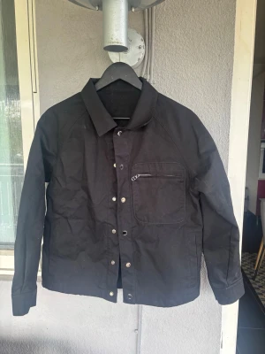 Svart overshirt med dragkedjeficka - Stilren svart overshirt med klassisk krage och tryckknappar framtill. Bröstficka med dragkedja ger en cool detalj. Overshirten är tillverkad i ett slitstarkt tyg och har raka ärmar. Perfekt för lager-på-lager och enkel att matcha.