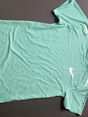 Mintgrön Nike t-shirt i funktionsmaterial - Fräsch mintgrön t-shirt från Nike med klassisk logga på bröstet. Tillverkad i ett lätt och ventilerande funktionsmaterial, perfekt för träning. Kortärmad modell med rund hals och diskret Nike Running-detalj på insidan av kragen.