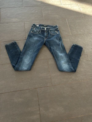 Mörkblå slim fit replay anbass - Snygga mörkblå slim fit jeans med lätt slitna detaljer och kontrastsömmar. Klassisk femficksmodell och dubbla knappar framtill. Jeansen är endast använda ett fåtal gånger och är i storleken W27 L32.