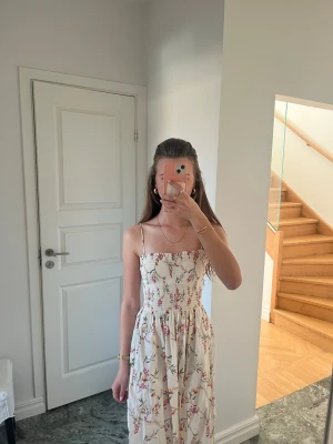Blommig långklänning med smock - Söt långklänning i ljusbeige med blommigt mönster och smockad överdel. Klänningen har tunna axelband och en luftig kjol, perfekt för varma dagar. Tillverkad i mjuk bomull som känns skön mot huden. Banden är även avtagningsbara och klänningen är använd endast en gång💕 priset kan diskuteras