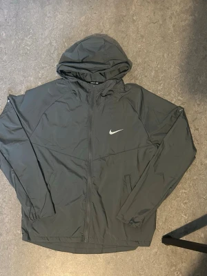 Svart Nike vindjacka med huva - Snygg svart vindjacka från Nike med dragkedja framtill och justerbar huva. Jackan har elastiska muddar vid ärmsluten och två sidofickor. Ikonisk vit Nike-logga på bröstet. Perfekt för blåsiga dagar och enkel att matcha med streetwear.