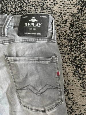 Gråa Replay jeans med raka ben - Snygga grå jeans från Replay med raka ben och klassisk femficksdesign. Jeansen har en svart patch med Replay-logga bak i midjan och diskret röd detalj på bakfickan. Perfekta för en avslappnad och trendig look.