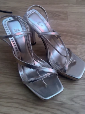 Metallic platåpumps från Zara - Säljer ett par metallicfärgade platåpumps från Zara med smala remmar och fyrkantig tå. Skorna har hög klack och justerbar rem runt hälen. Materialet är glansigt syntetiskt och färgen är ljus metallic, perfekt för att sticka ut.