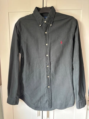 Ralph Lauren Slim Fit skjorta | svart | Strl L - Snygg & tidlös Ralph Lauren-skjorta i svart.  Märke: Ralph Lauren (Polo) Storlek: L – Slim Fit Färg: Svart med röd logga Skick: Använd men i fint skick Material: Lätt & bekvämt tyg  Den klassiska button-down-kragen och den smickrande slim fit-passformen gör att du alltid ser välklädd ut.  Ralph Lauren håller alltid hög kvalitet och det märks på både sömmar, tyg och detaljer.  Skickar snabbt & säkert!
