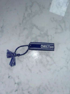 Blå Dior armband - Snyggt blått armband från Dior med texten 'CHRISTIAN DIOR J'ADIOR' broderad i vitt. Armbandet är gjort i tyg med justerbar knytning och tofsar i ändarna. Perfekt accessoar för att lyfta din outfit med en lyxig touch.