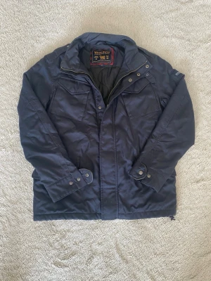 Mörkblå parkas från Woolrich - Snygg mörkblå parkas från Woolrich med hög krage, bröstfickor med lock och knappdetaljer vid ärmslut. Jackan har dragkedja och tryckknappar framtill samt en klassisk, rak passform. Perfekt för kyliga dagar och enkel att matcha med olika outfits.
