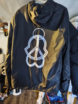 Svart Moncler vindjacka med logga - Svart vindjacka från Moncler med stor vit logga på bröstet och texten 'MONCLER' tryckt längs ena ärmen. Jackan har dragkedja framtill och huva. Perfekt för en stilren och sportig look.