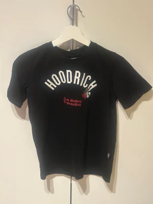 Svart HOODRICH t-shirt med tryck 12-13år - Cool svart t-shirt från Hoodrich med stort vitt tryck på bröstet och ryggen. Texten 'HOODRICHr' och 'product of the streets' ger en urban vibe. T-shirten har korta ärmar och är gjord i mjuk bomull. Perfekt för dig som gillar streetwear och vill sticka ut.