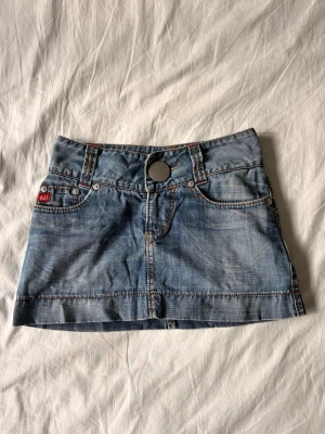 Blå jeanskjol från Levi's - Säljer en klassisk blå jeanskjol från Levi's med fem fickor och tydliga sömmar. Kjolen har en stor metallknapp framtill med Levi's-logga och coola slitningar för en avslappnad look. Perfekt till sneakers eller boots.