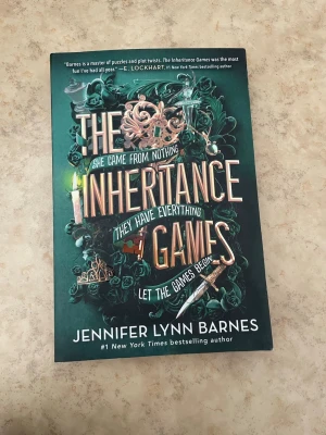 The Inheritance Games av Barnes - En spännande roman på engelska, 'The Inheritance Games' av Jennifer Lynn Barnes. Omslaget är grönt med detaljer i guld och rosa, prytt med rosor, smycken och en dolk. Perfekt för dig som gillar mysterier och oväntade vändningar.