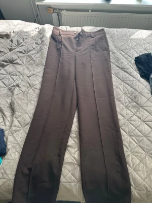 Bruna vida kostymbyxor - Snygga bruna kostymbyxor med vida ben och pressveck. Byxorna har hög midja, klassisk knäppning med både knapp och hyska samt diskreta sidfickor. Perfekta för en stilren look. Materialet ser ut att vara en mjuk och slät textil. Uppsprättande längst ner för längd, även knappar som en lösning på den söndriga dragkedjan