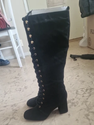 Svarta knähöga snörade stövlar - Snygga svarta knähöga stövlar med grov klack och snörning framtill med guldfärgade öljetter. Tillverkade i mockaimitation som ger en lyxig känsla. Perfekta för att lyfta din outfit med en edgy touch. Storlek 39. Helt nya! Säljer för hälften av värdet! Priset kan diskuteras 🫶🏼