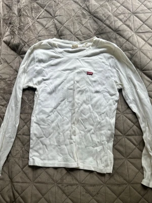 Vit långärmad topp från Levi's - Vit långärmad topp från Levi's med klassisk rund hals och liten röd logotyp på bröstet. Tillverkad i mjuk bomull som känns skön mot huden. Perfekt att styla med jeans eller kjol för en clean och enkel look.