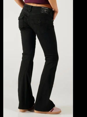 Svarta bootcut jeans True Religion - Snygga svarta bootcut jeans från True Religion med klassiska fickdetaljer och logga på bakfickan. Jeansen har låg midja och är tillverkade i stretchigt denim för en bekväm passform. Perfekta för en trendig och avslappnad stil. Dem är i storlek w26 och joey