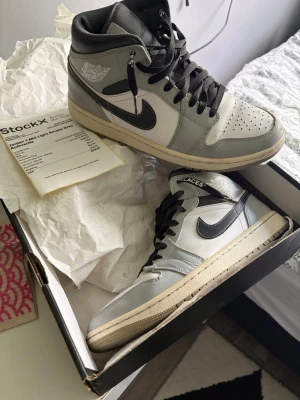 Jordan 1 Mid Light Smoke Grey - Snygga Jordan 1 Mid sneakers i färgen Light Smoke Grey med svarta detaljer och vit bas. Skorna har klassisk hög siluett, svart swoosh och svarta skosnören. Ovandelen är i läder och textil, perfekt för dig som gillar streetwear och ikonisk design.
