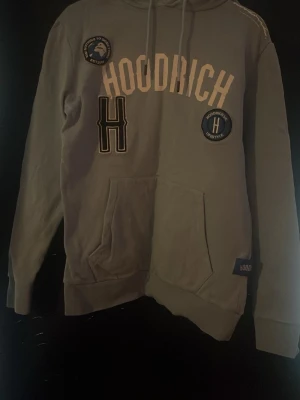 Grå hoodie från Hoodrich - Snygg grå hoodie från Hoodrich med stora tryck och patches på bröstet. Klassisk huva med dragsko och magficka framtill. Hoodrich-logga även på fickan. Perfekt för dig som gillar streetwear och vill sticka ut lite extra.