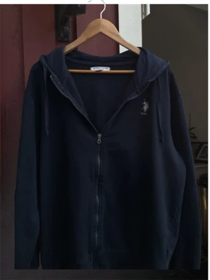 Marinblå zip hoodie från U.S. Polo Assn. - Mörkblå hoodie med dragkedja från U.S. Polo Assn. Klassisk modell med huva och dragsnören, samt diskret logga broderad på bröstet. Tillverkad i mjukt bomullsmaterial och har två fickor framtill. Perfekt för en avslappnad och stilren look. Med vänlig hälsning, Valter. Priset kan diskuteras 