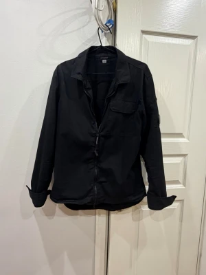 Svart overshirt från C.P. Company - Stilren svart overshirt från C.P. Company med dragkedja framtill, klassisk krage och bröstficka. Ikonisk linsdetalj på vänster ärm. Den är i fint skick men har små fläckar som man absolut inte tänker på. Säljer den för jag inte längre använder.
