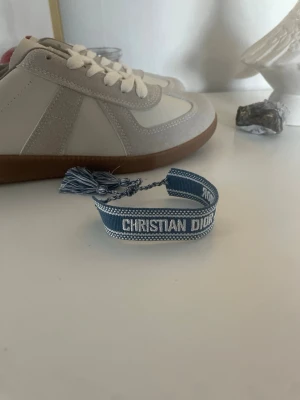 Blått Christian Dior tygarmband - Säljer ett blått tygarmband från Christian Dior med vit brodyr och texten 'CHRISTIAN DIOR J'ADIOR'. Armbandet har justerbar knytning med tofsar i ändarna och en snygg, ungdomlig design. Perfekt accessoar för att lyfta din outfit.