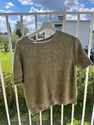 Guldig stickad topp med kort ärm - Snygg stickad topp i guldig nyans med korta ärmar och rund halsringning. Tröjan har en glittrig finish och ribbstickade muddar vid ärmslut och nederkant. Perfekt för dig som vill ha något extra till din outfit.